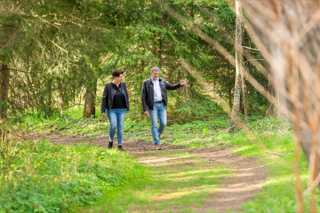 2 Personen, 1 Mann und 1 Frauen gehen durch einen Waldweg spazieren und unterhalten sich angeregt. Der Coach scheint ihr den Weg zu zeigen. Freundlich, einfühlsam und im geschützten Raum.