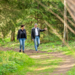 2 Personen, 1 Mann und 1 Frauen gehen durch einen Waldweg spazieren und unterhalten sich angeregt. Der Coach scheint ihr den Weg zu zeigen. Freundlich, einfühlsam und im geschützten Raum.