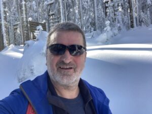 Christian, Dein Zuhörer steht im Schnee vor einem Wald und lächelt in die Kamera mit einer Sonnenbrille. Denn die Sonne scheint.