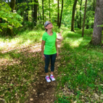 Eine Frau steht auf einem sonnigen Waldweg und blickt hinauf in die Baumkronen.