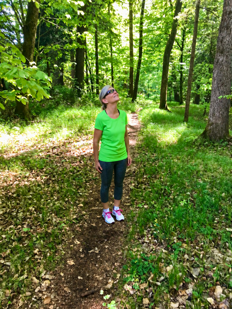 Eine Frau steht auf einem sonnigen Waldweg und blickt hinauf in die Baumkronen.
